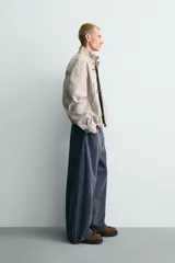 Cazadora relaxed fit confeccionada en tejido de algodón de color beige, con cuello subido y manga larga. Cuenta con bolsillos de plastrón en la cadera, bajo elástico y cierre frontal con cremallera oculta y alamares metálicos.
