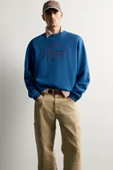 Sudadera azul con corte relaxed fit, cuello redondo y manga larga, confeccionada en tejido de algodón. Presenta un bordado frontal a contraste en color rojo con diseño ornamental y texto.