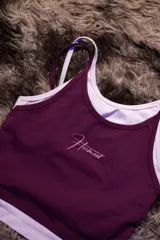 Top deportivo ajustado color morado oscuro con doble capa de tela y tirantes finos blancos. Presenta un logo manuscrito en tono rosado claro en el pecho.
