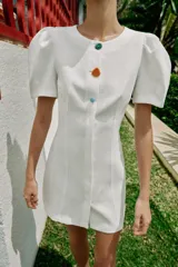 Vestido corto de color blanco con cuello redondo y mangas cortas abullonadas. Presenta un cierre frontal con botones decorativos de colores.