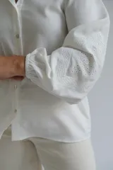 Camisa blanca de corte holgado con cuello clásico, cierre frontal de botones y mangas tres cuartos con sutil detalle bordado en los puños.