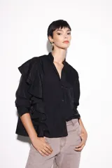 Camisa negra de cuello mao, de manga larga, con un gran volado plisado asimétrico que recorre desde el hombro derecho hasta el dobladillo inferior.