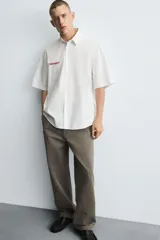 Camisa de algodón blanca con finas rayas verticales negras, corte relajado (relaxed fit) y manga corta. Presenta un bordado en hilo rojo en el lado izquierdo del pecho.