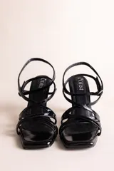 Sandalias de taco alto con tiras cruzadas en charol negro, puntera cuadrada y taco copa. Cuenta con tira ajustable en el tobillo con hebilla metálica.