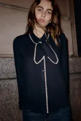 Blusa de color negro con cuello solapa, lazo frontal y manga larga. Presenta detalles de bordados en color crema en los bordes del cuello, el lazo, la tapeta de botones y los puños. Cierre frontal con botones.