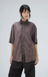 Camisa de corte oversize y mangas cortas, confeccionada en tela con efecto de teñido artesanal maltinto en tono gris. Presenta cuello clásico, cierre frontal con botones y costuras verticales decorativas en el frente.