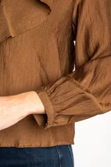 Blusa de color marrón con acabado satinado, cuello en V con volados y lazo de ajuste frontal. Presenta mangas largas con puños elásticos y un corte holgado.