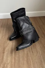 Bota de caña media confeccionada en cuero vacuno negro, con diseño de solapa superior y detalle de tachas metálicas plateadas en la parte central. Presenta punta cuadrada y taco bajo.