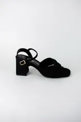 Sandalia de tacón cuadrado medio, confeccionada en material textil tipo gamuza color negro. Presenta un diseño de tiras cruzadas con nudo en la parte delantera y una tira ajustable con hebilla metálica en el tobillo.