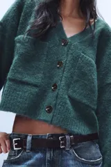 Chaqueta de punto de textura suave en color marrón oscuro, con escote en pico, manga larga y cierre frontal con botones. Presenta dos bolsillos de plastrón en la parte delantera.