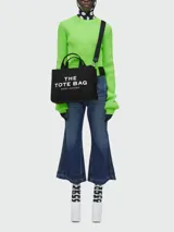 Bolso tote de lona color verde oliva con estampado frontal en blanco que dice 'THE TOTE BAG MARC JACOBS'. Cuenta con doble asa superior, correa de hombro ajustable y extraíble en color negro, y cierre superior con cremallera.