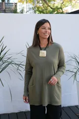 Sweater de punto color verde oliva, con cuello redondo, mangas largas y bolsillos frontales tipo parche. Presenta un corte holgado y largo, con terminaciones acanaladas en el ruedo y los puños.