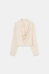 Camisa corta de manga larga, color crema o beige muy claro, con cuello solapa y escote drapeado o fluido tipo cascada.