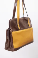 Cartera de hombro tipo portafolio confeccionada en cuero vacuno, con diseño estructurado que combina marrón liso y un panel frontal en cuero texturizado color mostaza. Posee doble asa larga y cierre superior.