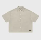 Sobre camisa de gabardina color beige, con corte boxy fit, cuello clásico, cierre frontal con botones y etiqueta decorativa en el ruedo inferior.