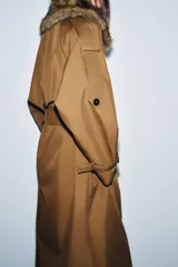 Trench largo color marrón, de corte amplio, con cuello solapa desmontable de pelo sintético marrón. Presenta cierre cruzado con botones oscuros y cinturón de tela anudado a la cintura.