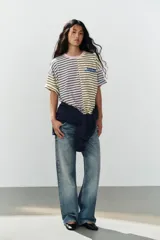 Camiseta oversize con diseño bicolor a rayas horizontales, combinando un lado en tonos rosado y verde oscuro, y el otro en beige y marrón. Cuenta con cuello redondo, mangas cortas amplias y texto bordado en azul en el pecho.