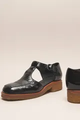 Zapato tipo guillermina de cuero charol color marrón oscuro, con tira T y hebilla ajustable en el lateral. Posee puntera cuadrada y suela de goma color caramelo con taco bajo.