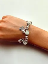 Pulsera plateada con dijes colgantes, incluyendo un trébol, un corazón, esferas de color verde claro y un dije transparente, con estructura de eslabones y cuentas decorativas.