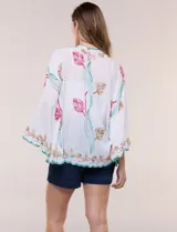Kimono de poliéster blanco con diseño abierto, mangas amplias y bordados coloridos de motivos florales y arabescos, con terminaciones en ondas de hilo verde.