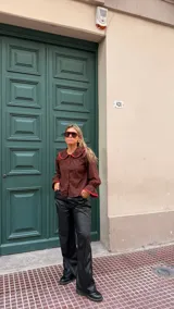 Camisa de color marrón chocolate con cuello Peter Pan y puños con volados, ambos detalles resaltados con un ribete en color rojo. Presenta cierre frontal con botones rojos.