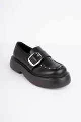 Mocasín negro de plataforma con suela chunky, tira frontal con hebilla metálica plateada y detalles de tachas en el borde del empeine.
