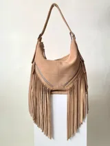 Bolso tipo hobo color tostado, hecho de cuero con flecos largos en el frente y dos bolsillos con cierre. Tiene una manija regulable con hebillas metálicas.