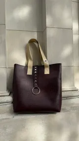 Bolso tote color borgoña con textura símil piel de cocodrilo, doble asa corta color beige y cierre superior. Presenta una tira vertical con apliques metálicos y un aro plateado decorativo.