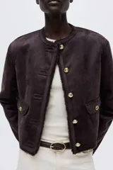 Chaqueta corta de gamuza sintética en color marrón oscuro, con forro interior de peluche. Presenta cuello redondo, cierre frontal con botones metálicos dorados y dos bolsillos delanteros con solapa y botón.