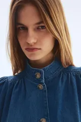 Campera de sarga de mezclilla azul, con cuello elevado, cierre frontal de botones y canesú fruncido. Tiene mangas largas abullonadas con puños ajustados y bolsillos laterales insertados.
