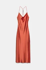 Vestido midi satinado de color naranja tostado, con escote fluido tipo cowl y tirantes finos cruzados en la espalda con detalle de aplicación metálica. Presenta un diseño largo con tajo lateral en la falda.