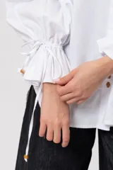 Camisa de algodón blanca de silueta amplia, con cuello camisero, botonadura frontal con botones decorativos, mangas abullonadas extragrandes y detalles de tiritas ajustables con cuentas de resina en los puños.
