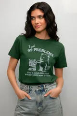 Remera de algodón color verde con cuello redondo y mangas cortas. Presenta un estampado frontal en color blanco con un oso, un carrito de compras y la frase 'I got 99 PROBLEMS And a sweet little treat WOULD SOLVE MOST OF THEM'.