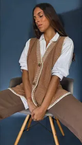 Chaleco tejido color tostado con destellos de lurex, escote en V y cierre frontal con botones. Presenta detalles de bordados y terminaciones en crochet en color crudo.