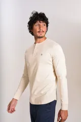 Remera de manga larga tipo Henley, color beige, con cuello redondo y tapeta con botones. Presenta un pequeño logo bordado en el pecho.