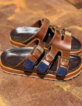 Sandalias de cuero marrón con tres tiras ajustables con hebillas doradas y detalles en rojo.