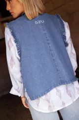 Chaleco de jean azul con diseño asimétrico, cierre lateral y detalle de volados en los bordes. Presenta un estilo rústico y moderno.