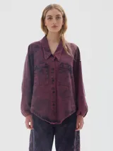 Camisa oversized de denim con lavado acid wash en tonos morados y azul oscuro. Presenta cuello camisero, escote en V, dos bolsillos frontales con tapa y botones oscuros. Las mangas son largas tipo balloon con puño ajustado. El bajo es irregular.