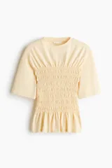 Remera de punto de algodón color amarillo claro, con cuello redondo, hombros levemente caídos, mangas cortas y cuerpo completamente fruncido con detalle de volados en el ruedo.
