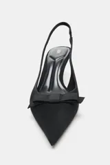 Zapato de tacón destalonado tipo slingback, confeccionado en tejido negro. Presenta punta fina, un pequeño lazo decorativo en la parte delantera y una tira elástica en el talón para mayor ajuste.