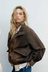 Chaqueta encerada color marrón con efecto lavado, cuello subido y manga larga. Cuenta con bolsillos frontales de plastrón y cierre de cremallera oculto por solapa con botones a presión. Los puños reversibles dejan ver un forro interior con estampado a cuadros.