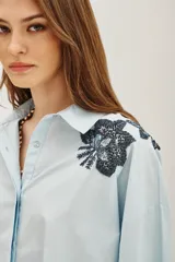 Camisa blanca de algodón con cuello camisero y mangas largas abullonadas con puños ajustados. Presenta detalles bordados en los hombros con motivos vegetales y aplicaciones brillantes.