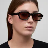 Lentes de sol con montura cuadrada de color negro y cristales polarizados en tono marrón. Presentan un diseño retro con parte superior plana y patillas con detalle sobresaliente.