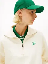 Sweater de felpa color blanco marfil, con cuello alto y cierre frontal de media cremallera. Presenta mangas largas con puños y dobladillo acanalados, y un pequeño logo script bordado en verde en el pecho.