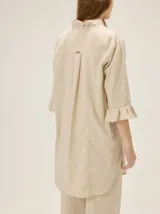 Camisa de lino de silueta relajada y largo irregular, color beige, con cuello mao, botones frontales a contratono y mangas tres cuartos terminadas en volados.