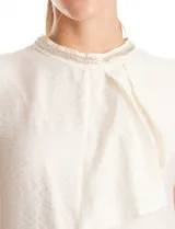 Blusa sin mangas color blanco, confeccionada en crepe con acabado satinado. Presenta un cuello alto decorado con mostacillas y una sobrecapa drapeada en el frente. La espalda cuenta con una abertura tipo gota y cierre de botón.
