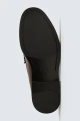 Mocasín de vestir color marrón oscuro, con parte superior lisa, detalle de antifaz con borlas en el empeine y horma redonda. Suela de goma a tono.