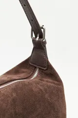 Cartera de gamuza negra con textura suave y acabado aterciopelado. Cuenta con una silueta amplia y flexible, cierre superior metálico, correa regulable y detalles laterales con hebillas.