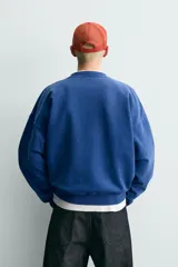 Sudadera de corte boxy fit en color azul con efecto lavado, cuello redondo y manga larga. Presenta estampado frontal con tipografía variada y gráficos estilo universitario. Interior afelpado y acabados en rib.