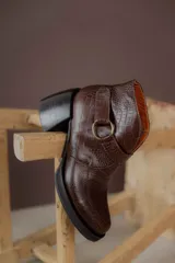 Bota corta de cuero vacuno con textura de reptil, color marrón oscuro. Cuenta con un taco cuadrado negro, suela de plataforma y una tira decorativa con hebilla metálica circular en el tobillo.